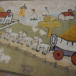 Close-up of a hooked rug showing a landscape pattern with white houses, green hills, and a wagon.