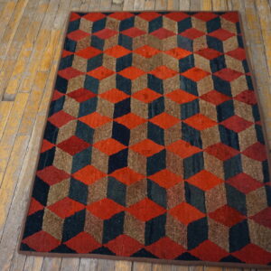 Low-pile rug with red, teal, and brown tumbling cubes rests on worn yellow wood flooring.
