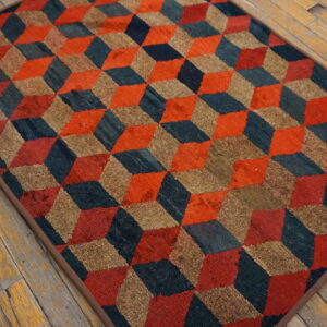 Textured rug with interlocking cubes pattern in orange, dark blue, and tan on weathered wood flooring.
