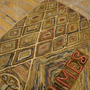 A corner of a brown and gold hooked rug with geometric diamonds and red lettering rests on aged wood flooring.