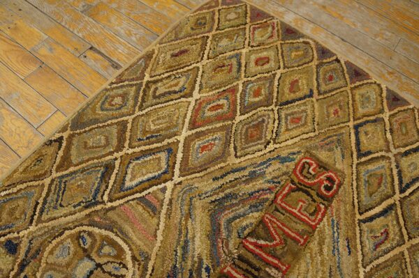 A corner of a brown and gold hooked rug with geometric diamonds and red lettering rests on aged wood flooring.