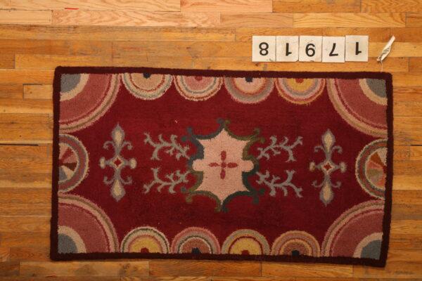 Deep red rug with stylized floral motifs and multicolored scalloped borders lies on hardwood flooring.