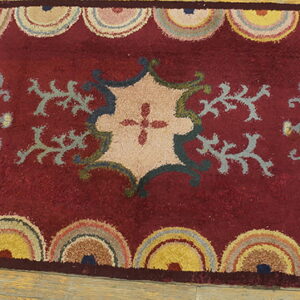 Rectangular maroon rug with a tan central motif and colorful scalloped borders on wood planks.