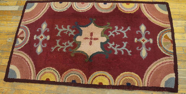 Rectangular maroon rug with a tan central motif and colorful scalloped borders on wood planks.