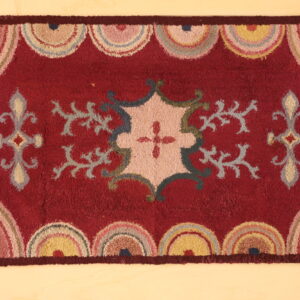 Textured red rug with a central pastel floral design and repeating multicolored scallop borders on a light floor.