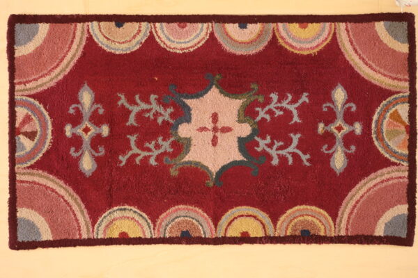 Textured red rug with a central pastel floral design and repeating multicolored scallop borders on a light floor.