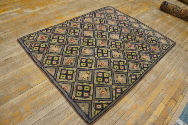 A grayish-blue hooked rug featuring a grid of flowers and geometric boxes rests on distressed yellow wood planks.