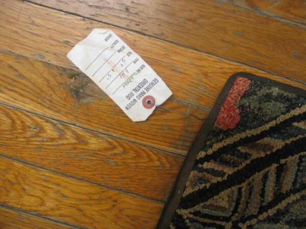 A textured rug corner featuring dark red, black, and green patterns rests on distressed natural wood plank flooring beside a paper tag.
