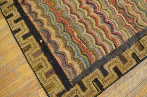 Close-up of a hooked rug featuring multicolored wavy stripes and a gold and black geometric greek key border on wood flooring.