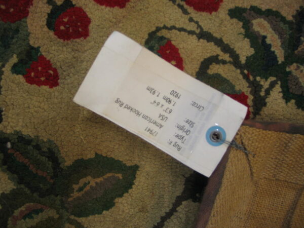 Textured rug with red floral elements and green foliage on a tan background, partially covered by a white tag.