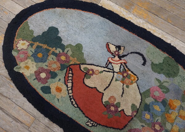 Hooked oval rug with a central female figure, red dress, and multicolored floral border on distressed wood floor.