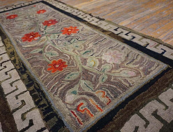 A hooked runner rug featuring red flowers, green vines, and a greek key border on wood floor.