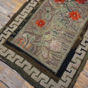 Highly textured hooked rug featuring red flowers and a Greek key border on weathered wood flooring.