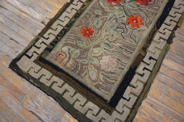 Highly textured hooked rug featuring red flowers and a greek key border on weathered wood flooring.