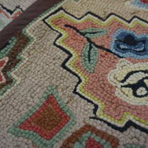 Close-up of a hooked rug texture showing pink, blue, beige, and green floral and geometric patterns.