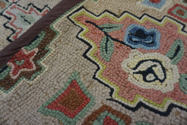 Close-up of a hooked rug texture showing pink, blue, beige, and green floral and geometric patterns.