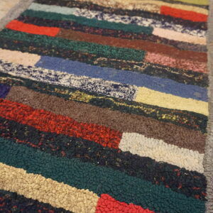 A close-up shows a chunky, multicolored striped rug with a thick gray border on a wooden floor.