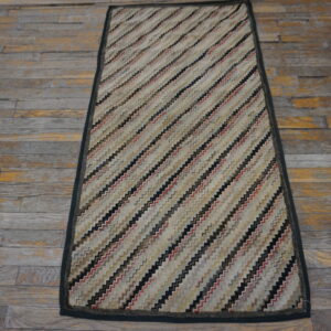 A rectangular low-pile rug with diagonal zigzag stripes in beige, black, and red rests on distressed wood flooring.