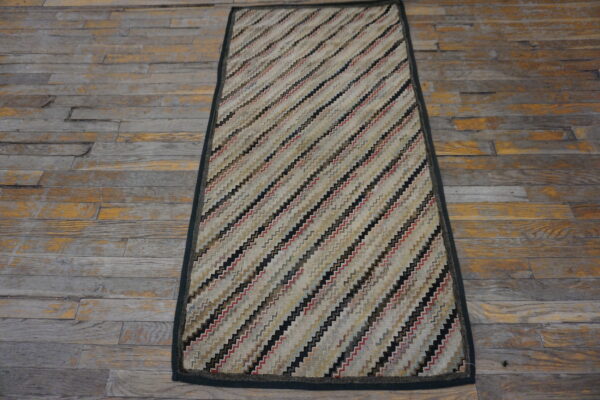 A rectangular low-pile rug with diagonal zigzag stripes in beige, black, and red rests on distressed wood flooring.