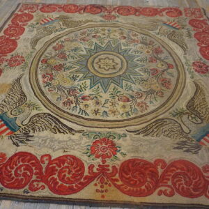 Textured beige rug with a blue star medallion, eagle motifs, and a broad red scrolling border on wood planks.