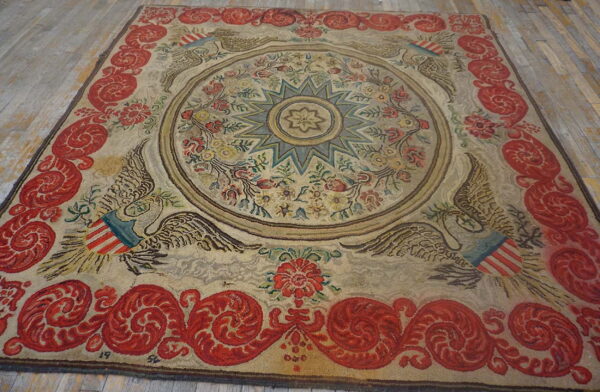 Textured beige rug with a blue star medallion, eagle motifs, and a broad red scrolling border on wood planks.