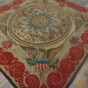 Beige rug with red scrolling borders and eagle motifs rests on worn wood flooring.