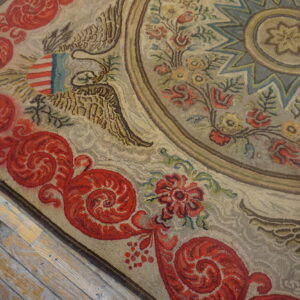 Textured beige and red hooked rug displaying an ornamental eagle design on worn hardwood floors.