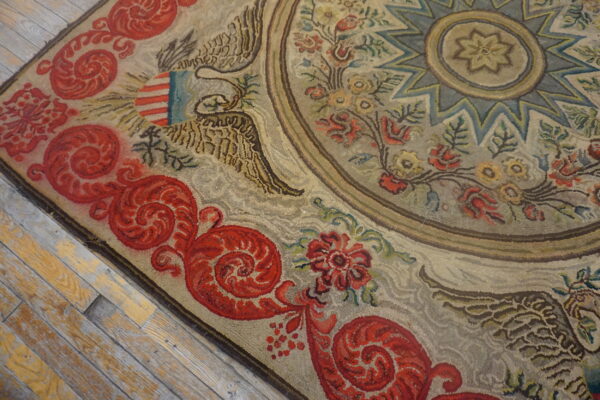 Textured beige and red hooked rug displaying an ornamental eagle design on worn hardwood floors.