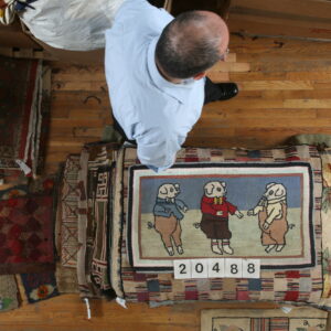 A man stands on a wood floor near a stack of textured rugs, one featuring three cartoon pigs.
