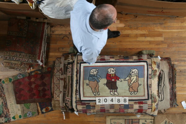 A man stands on a wood floor near a stack of textured rugs, one featuring three cartoon pigs.