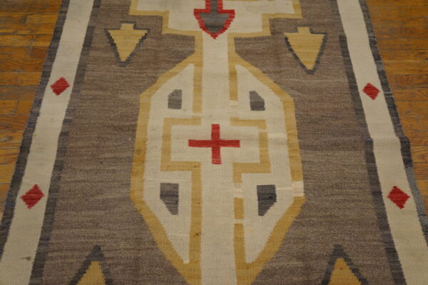 Brown flatwoven rug with gold and cream geometric patterns and red cross accent on wood floor.