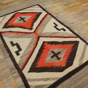 Geometric flatweave rug in orange, dark brown, cream, and gray rests on distressed plank wood flooring.