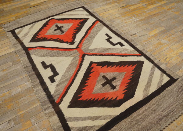 Geometric flatweave rug in orange, dark brown, cream, and gray rests on distressed plank wood flooring.