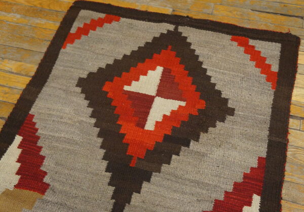 Gray flatweave rug with red, brown, and cream geometric stepped diamond design on wood planks.