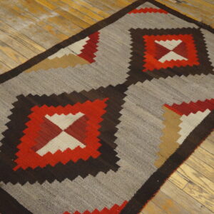 Geometric flatweave rug in red, brown, tan, and cream on a heather gray field, resting on worn wood planks.