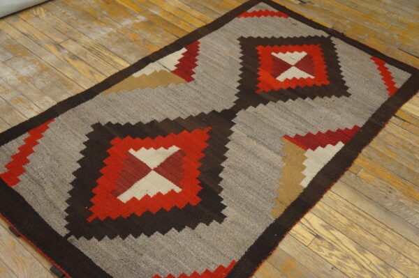 Geometric flatweave rug in red, brown, tan, and cream on a heather gray field, resting on worn wood planks.