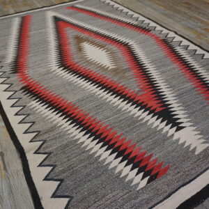 Gray flatweave rug with red, black, and cream serrated diamond patterns rests on distressed plank flooring.