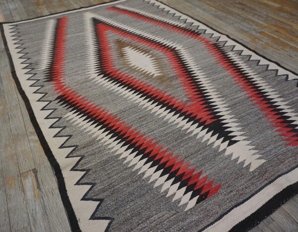Gray flatweave rug with red, black, and cream serrated diamond patterns rests on distressed plank flooring.