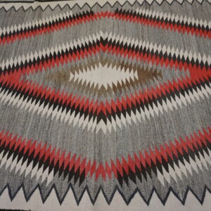 Geometric rug with a large serrated diamond motif using gray, red, black, brown, and cream colors.