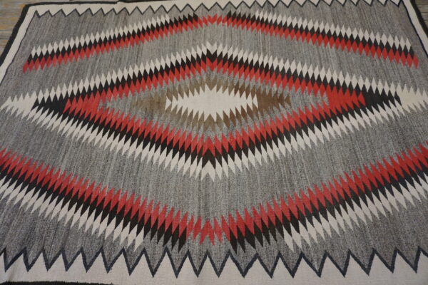Geometric rug with a large serrated diamond motif using gray, red, black, brown, and cream colors.