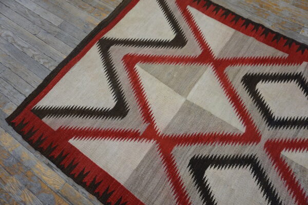 A flatweave rug with serrated geometric patterns in cream, red, gray, and dark brown on gray wood.