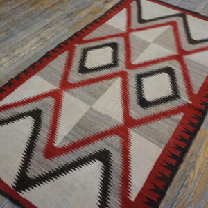 Red, black, gray, and cream flatwoven rug with large serrated geometric patterns on distressed wood flooring.