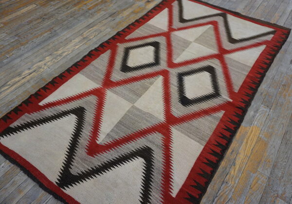 Red, black, gray, and cream flatwoven rug with large serrated geometric patterns on distressed wood flooring.