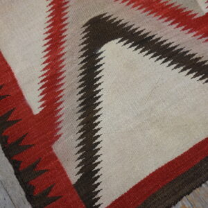 Geometric rug in cream, red, and dark brown with serrated edges lies on gray distressed wood flooring.