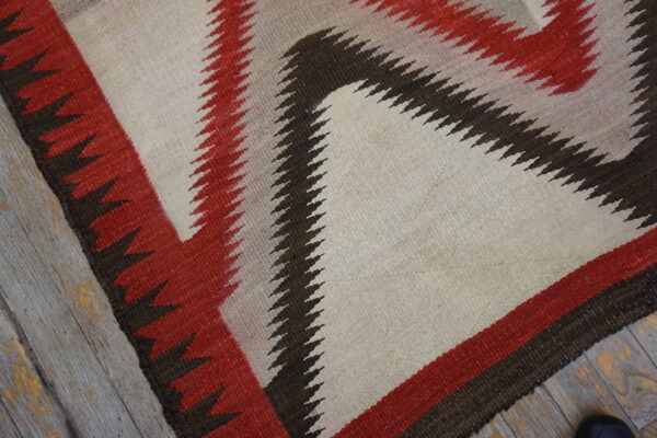 Geometric rug in cream, red, and dark brown with serrated edges lies on gray distressed wood flooring.