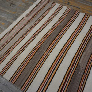 Striped flatweave rug featuring cream, gray, orange, red, and black on distressed gray plank flooring.