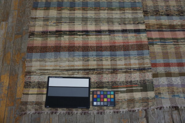 Textured flat-weave rug featuring variegated horizontal stripes and checkered blocks on rustic wood planks.