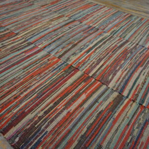 Multicolored flatweave rag rug with thin vertical stripes in reds, blues, and browns on worn wooden flooring.
