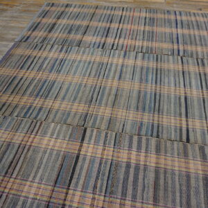 Muted gray, blue, and tan plaid rug assembled from woven panels, displayed on weathered wood planks.