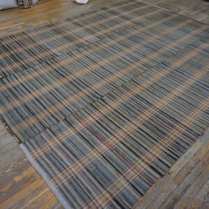 Flat-woven plaid rug with tan, gray, and blue stripes lying on rustic, multi-toned wood flooring.